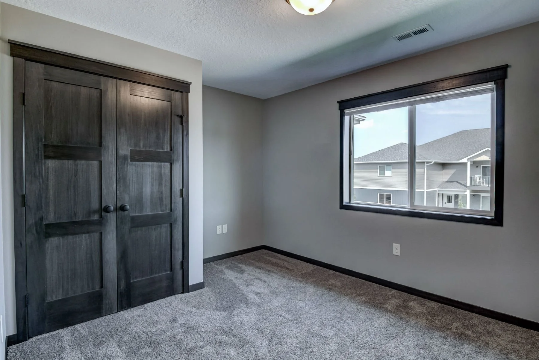 Empty bedroom with a window viewing neighboring houses, a dark wood closet with double doors, power outlets on the wall, and a ceiling light fixture.