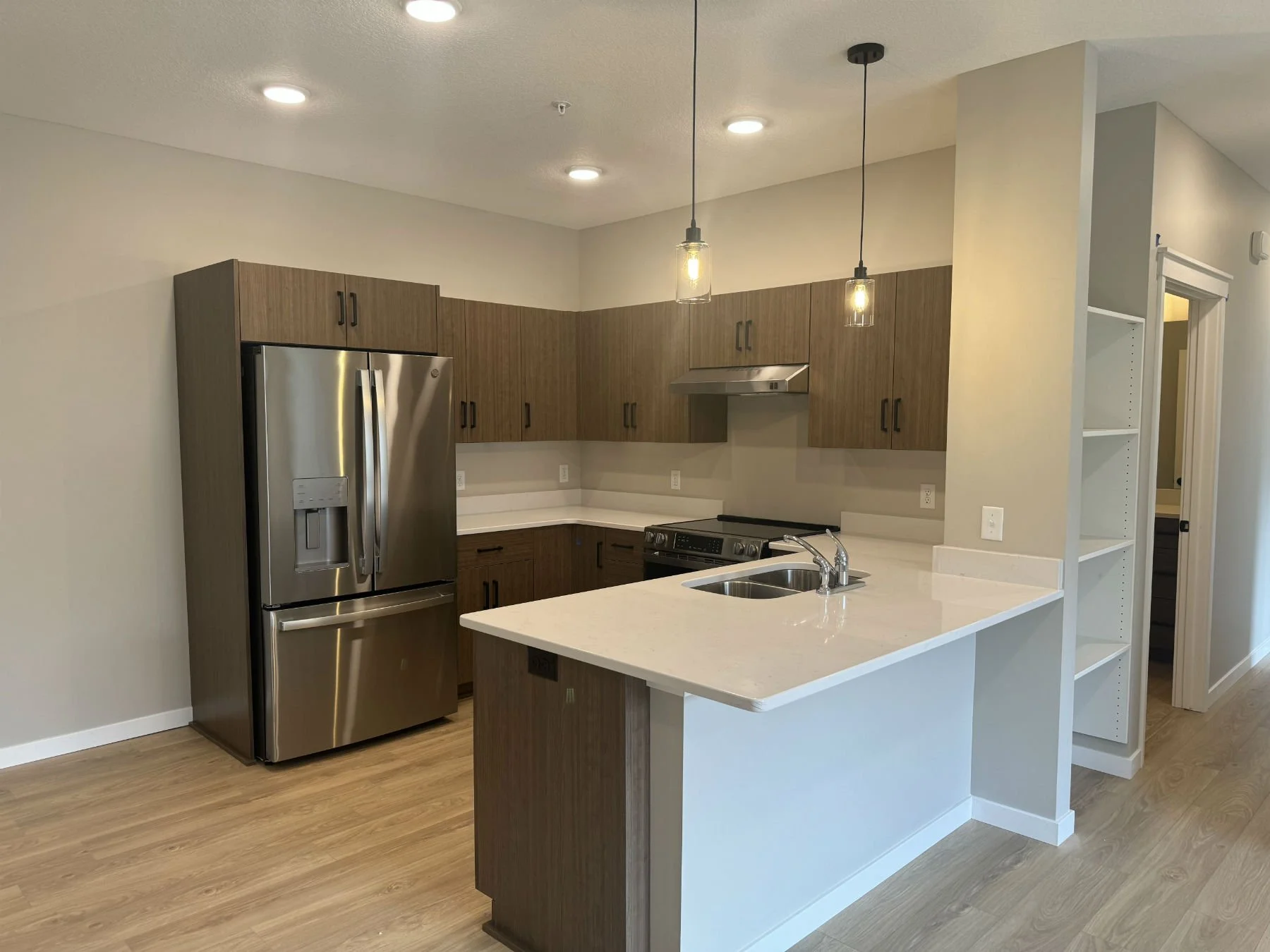 A modern kitchen with light-colored walls, wooden cabinets, stainless steel refrigerator, and a white countertop with a sink. There are hanging pendant lights above the countertop, a stove with an exhaust hood, and a small open shelving unit on the r