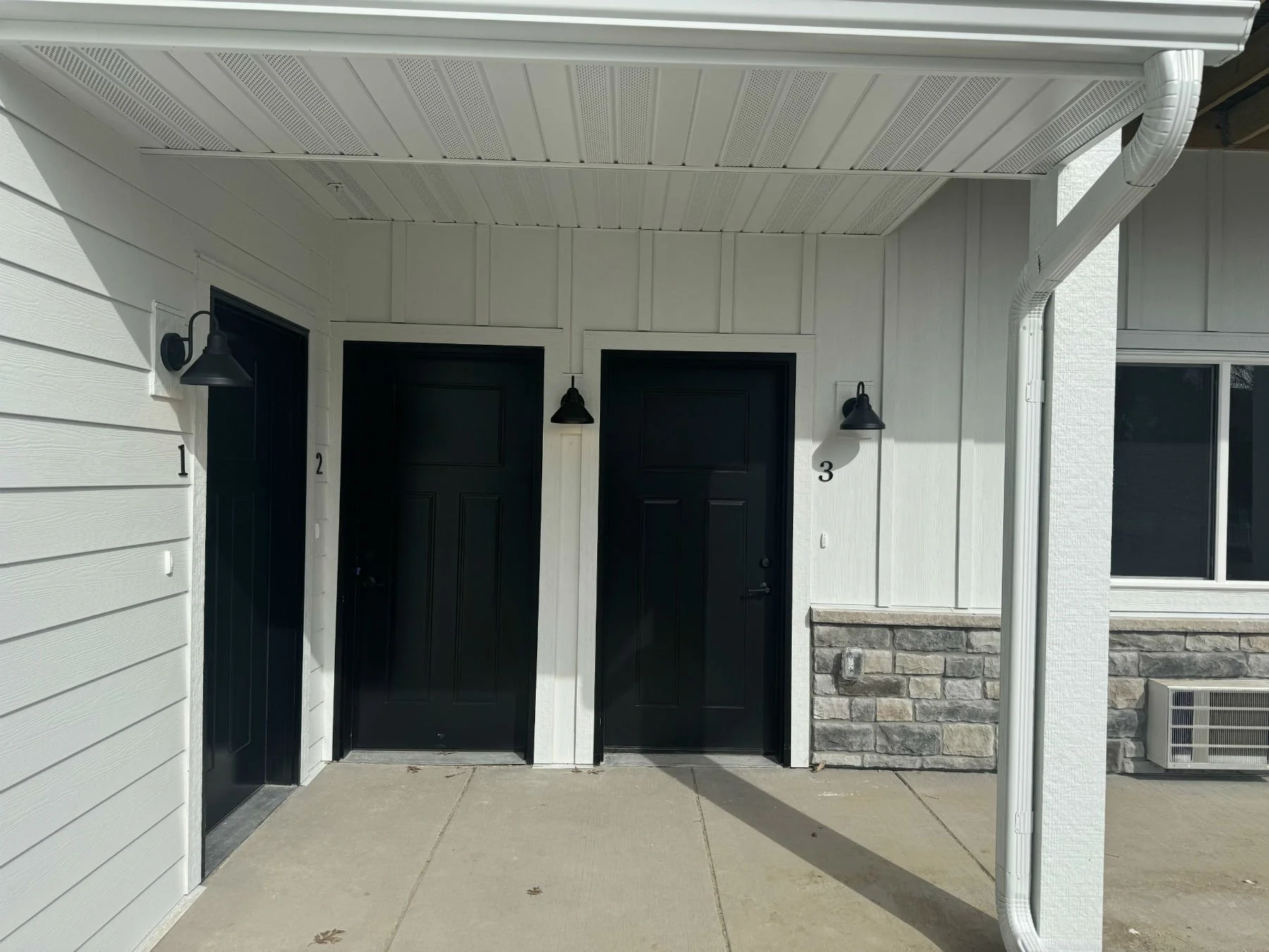 Front porch area with two black doors, wall-mounted exterior lights, white siding, stone accents, window, drainage pipes, and a concrete sidewalk.
