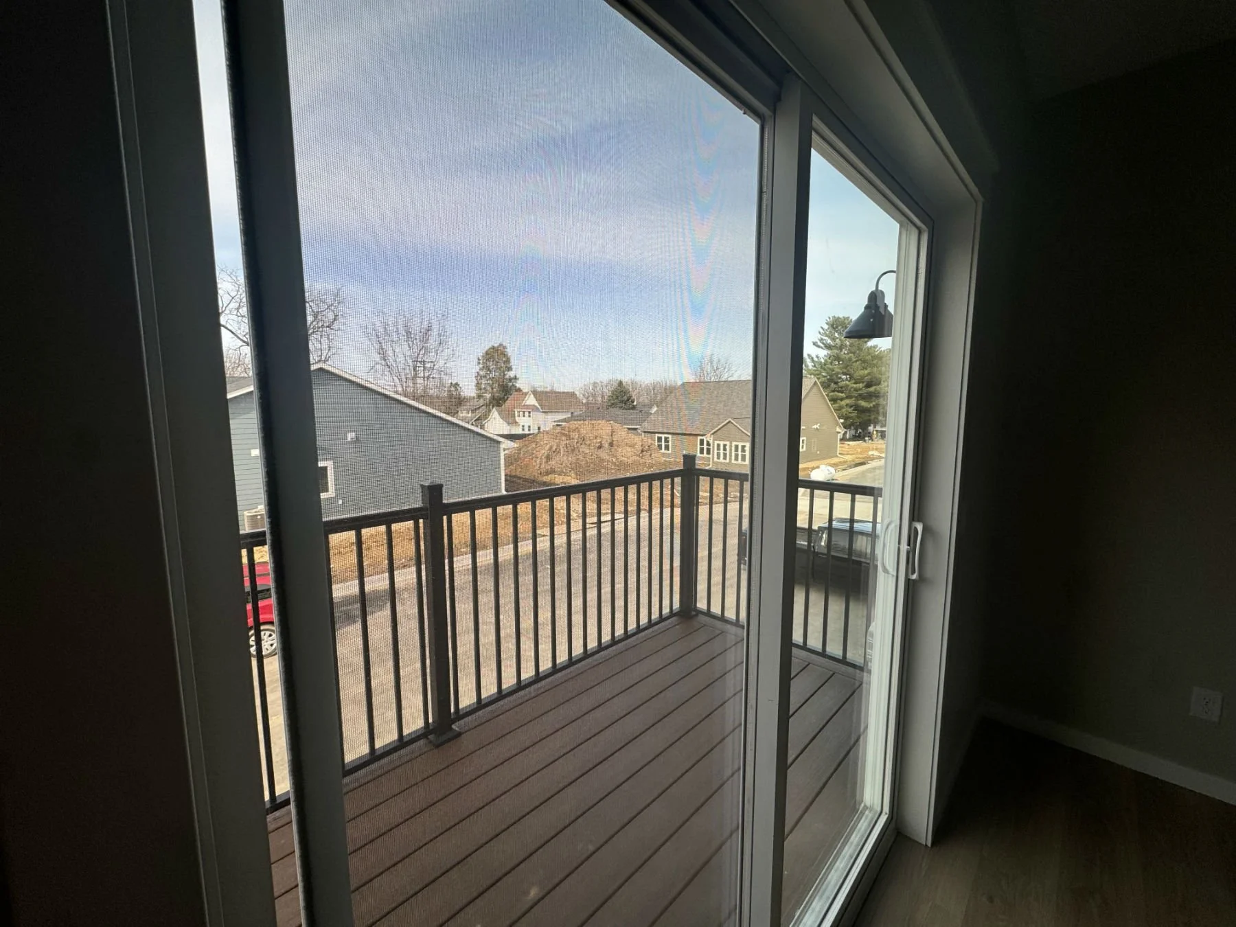 View from inside a room looking out through a sliding glass door to a small balcony with a black railing. Outside are residential houses, some trees, a parked car, and a partly cloudy sky.