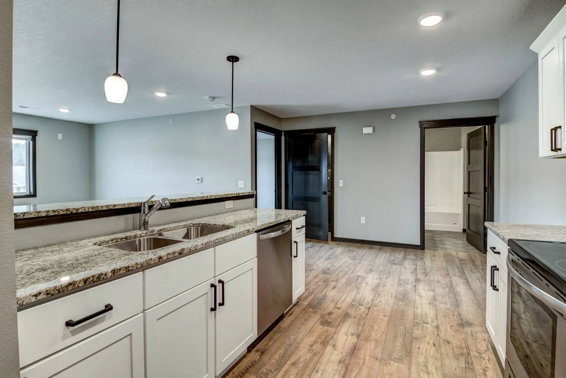 Modern kitchen with white cabinets, granite countertops, stainless steel sink, wooden flooring, and black-framed windows and doors. There is a view into a small room or bathroom with a bathtub.