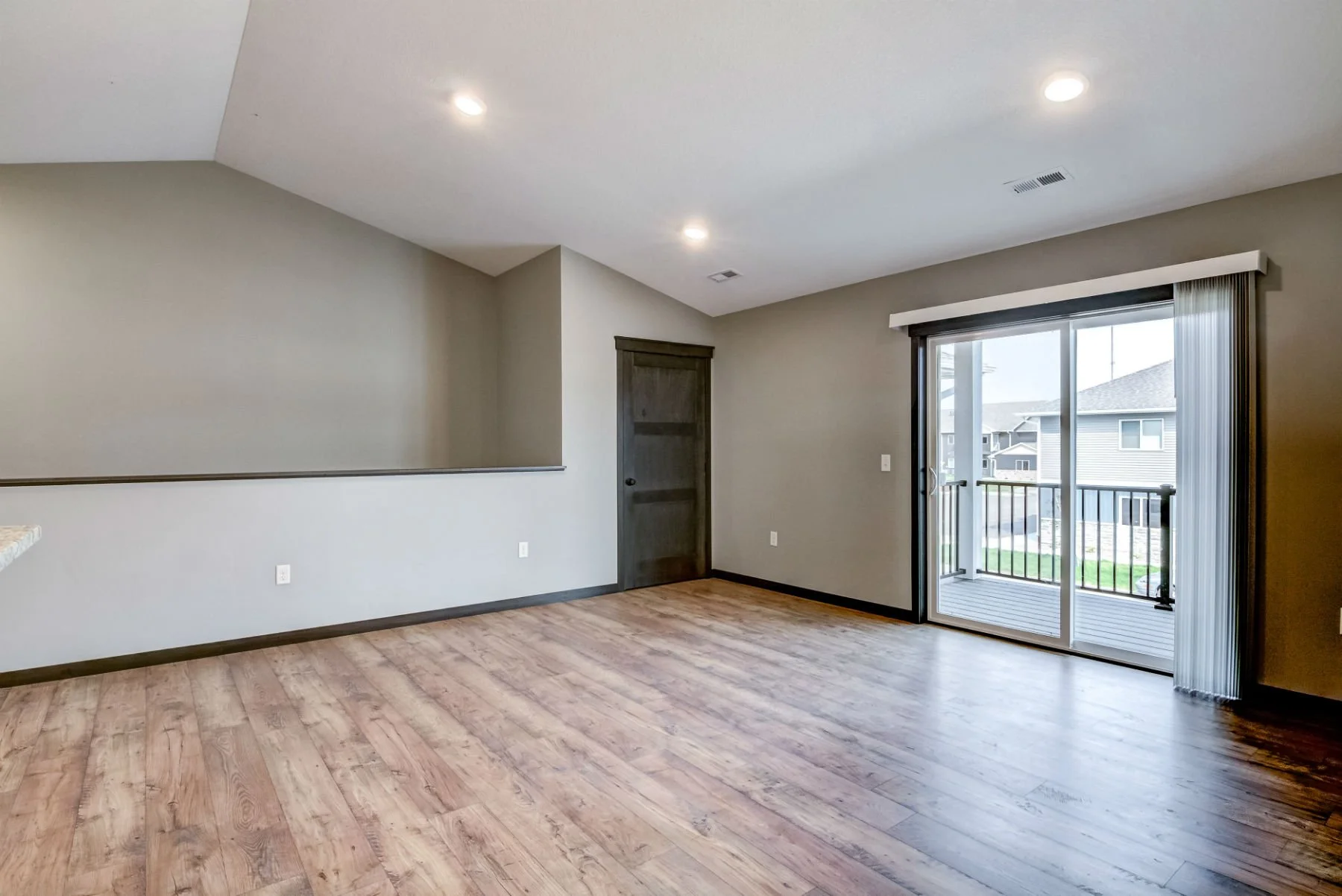 Empty living room with wood flooring, a sliding glass door leading to a balcony, and beige walls with a dark wooden door.