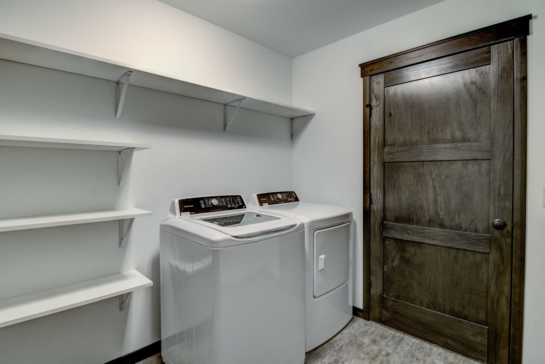 A laundry room with white wall shelves, a top-loading washing machine, a matching dryer, and a large dark wood sliding door.