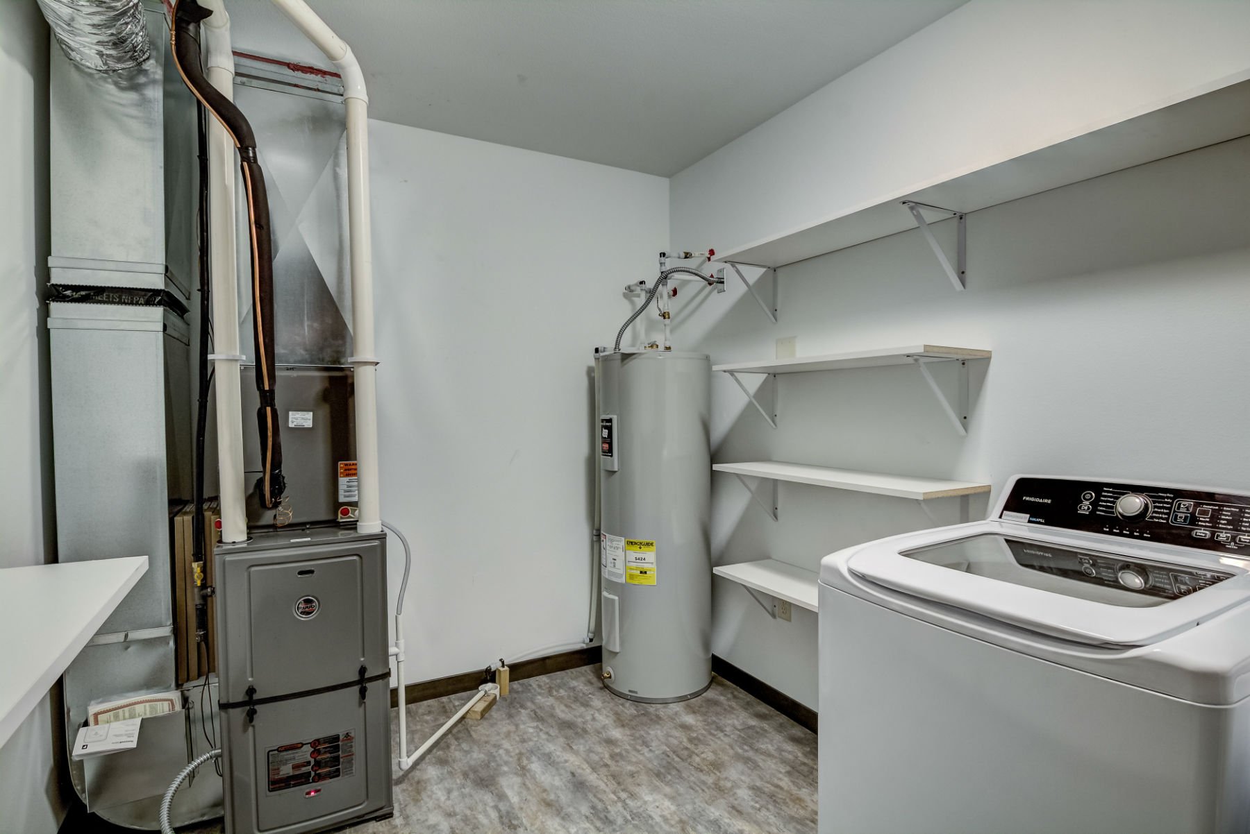 A laundry room with a washing machine, water heater, shelving, and utility pipes.