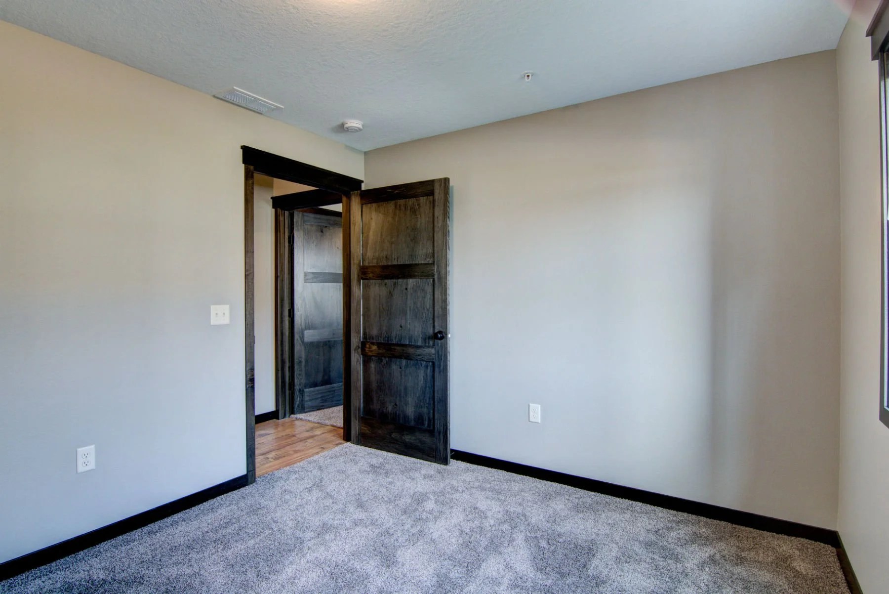 An empty room with light gray walls, dark brown trim, gray carpet, a partially open dark wood door leading to another room, and a window on the right side.