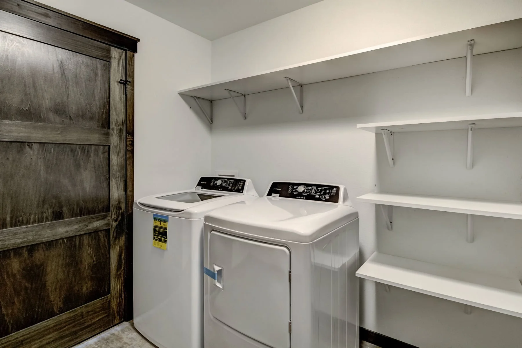 Laundry room with white washing machine and dryer, wooden sliding door, and white shelves on the wall.