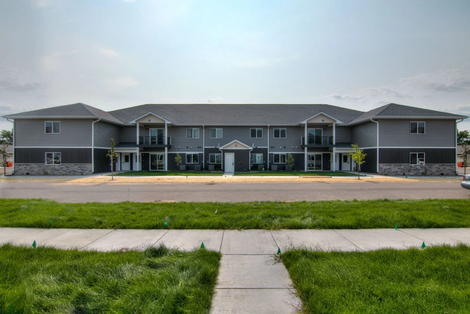 A modern, multi-unit apartment building with two floors, gray siding, and small balconies. The building has a brick and stone foundation, with small trees and a grassy area in front. The sky is partly cloudy.