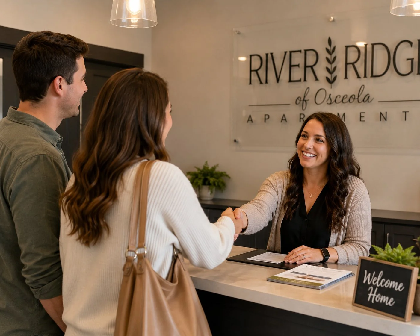 A woman greets a couple at a reception desk in an apartment complex lobby, shaking hands and smiling.