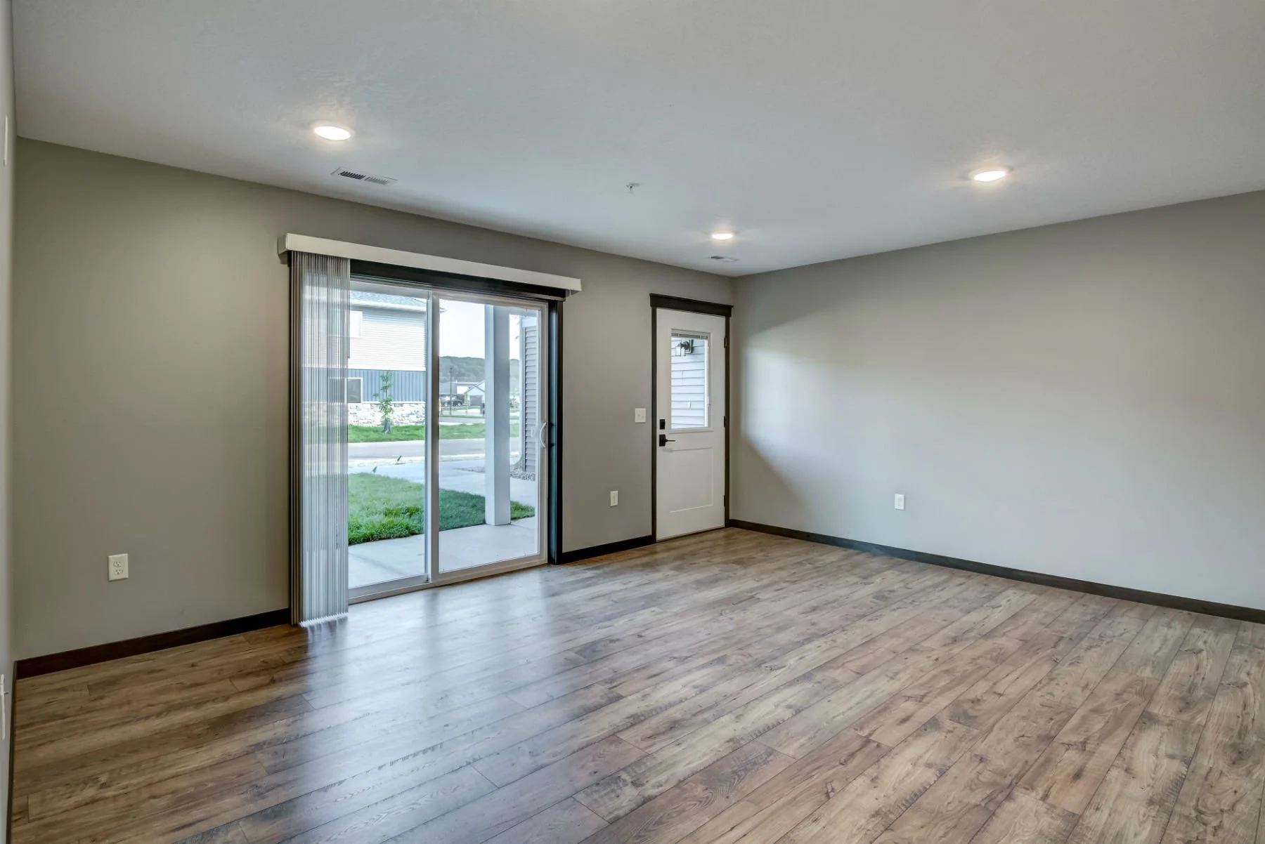 Empty living room with wood flooring, a sliding glass door with vertical blinds leading outside, a white door, and minimal wall decorations.