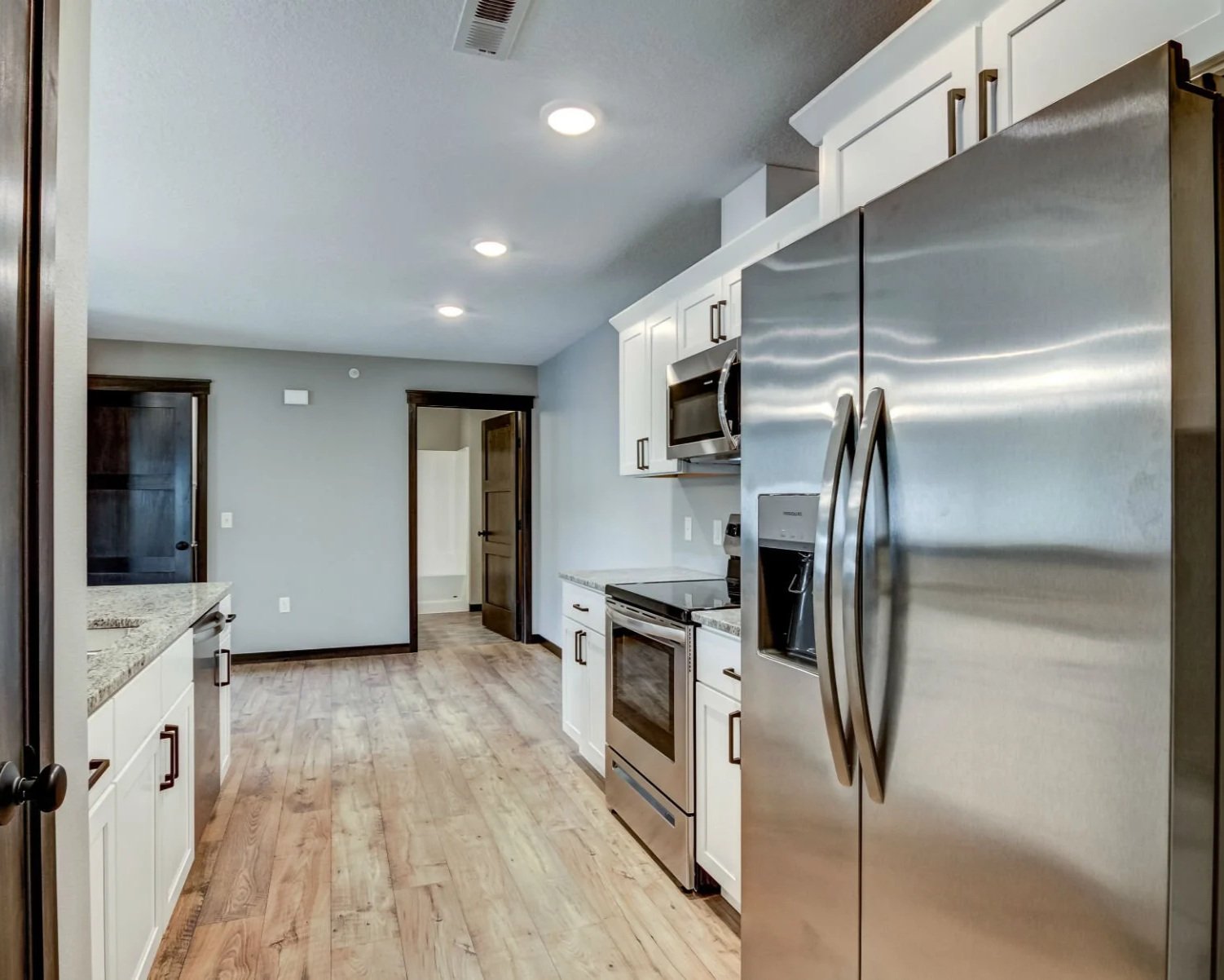 Modern kitchen with stainless steel side-by-side refrigerator, microwave, oven, and white cabinets with dark handles, hardwood flooring, and recessed lighting.