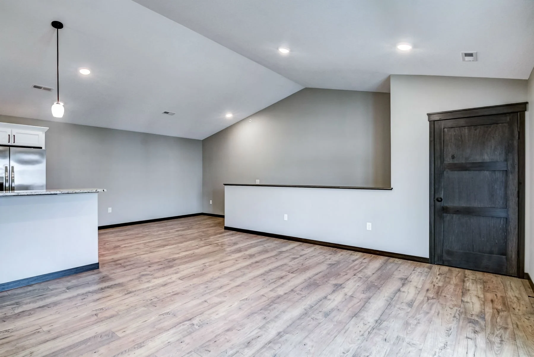 Empty room with wooden flooring, white walls, and a dark wooden door. Partially visible kitchen area with a granite countertop and stainless steel refrigerator. Recessed ceiling lights and a hanging light fixture.
