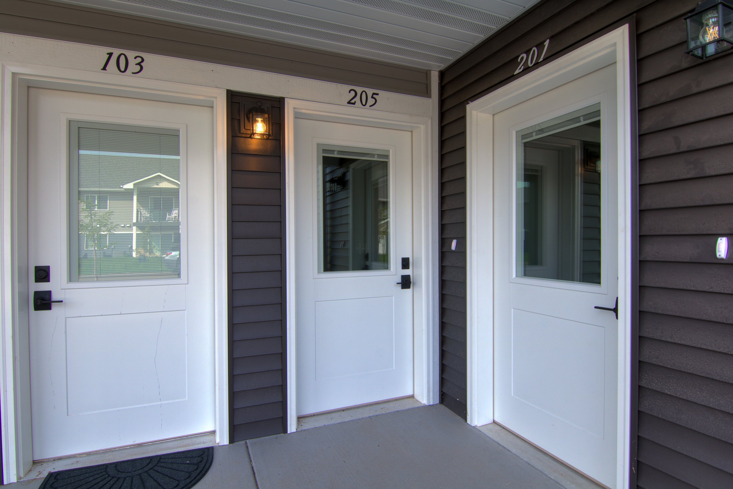 Entrance area showing two white doors with window panes, black door handles, and house numbers 103, 205, and 201. Side windows and wall-mounted lights are visible, with reflections of neighboring houses in the windows.
