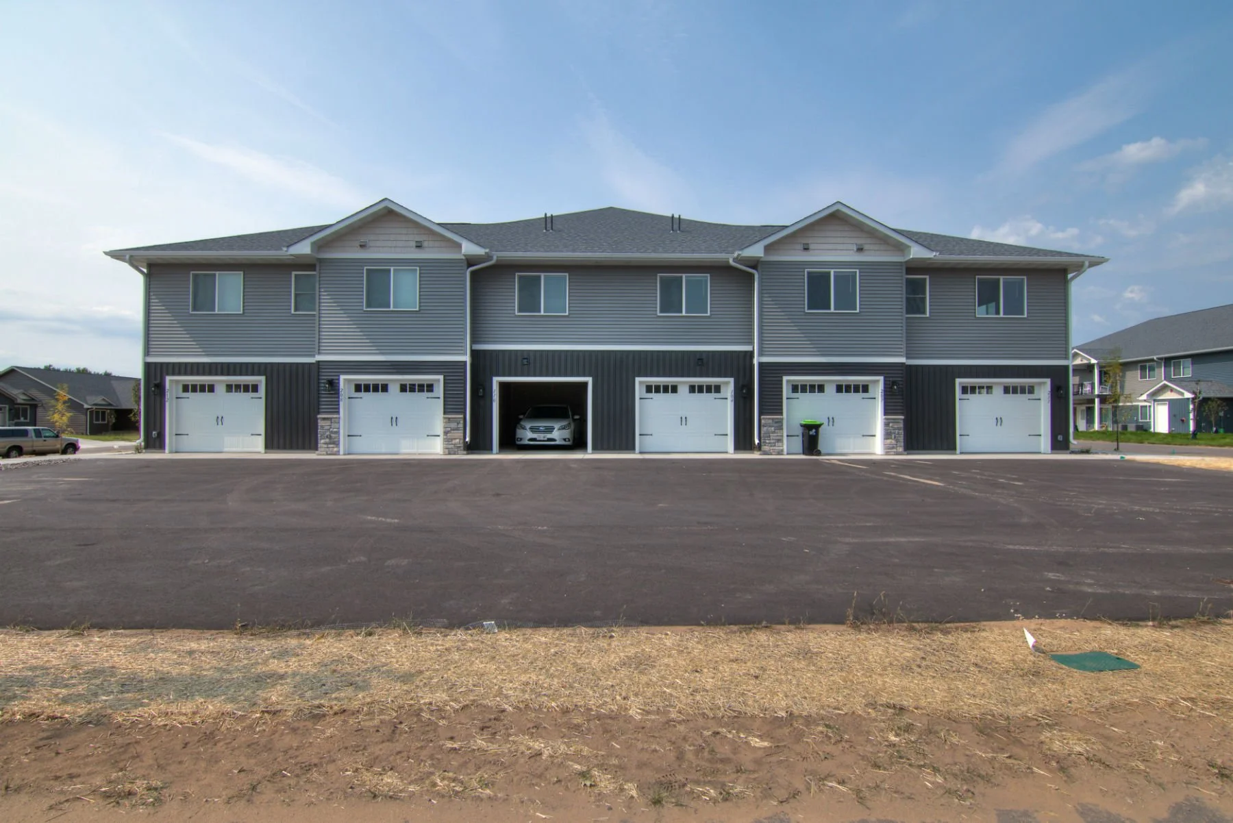 A modern multi-family apartment building with five garage units on the ground floor, one of which houses a white car. The building is gray with white trim and has two stories with multiple windows. The foreground features a paved parking area and a s