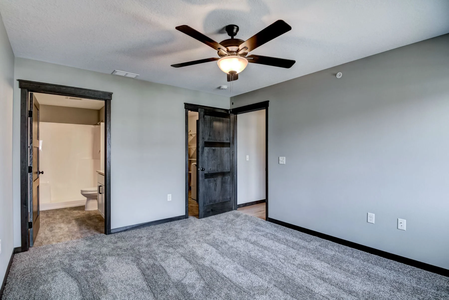 A bedroom with gray carpet, light gray walls, dark wood trim, and a ceiling fan with a light. There is an open door leading to a bathroom with a toilet visible, and a sliding dark wood door leading to a closet.