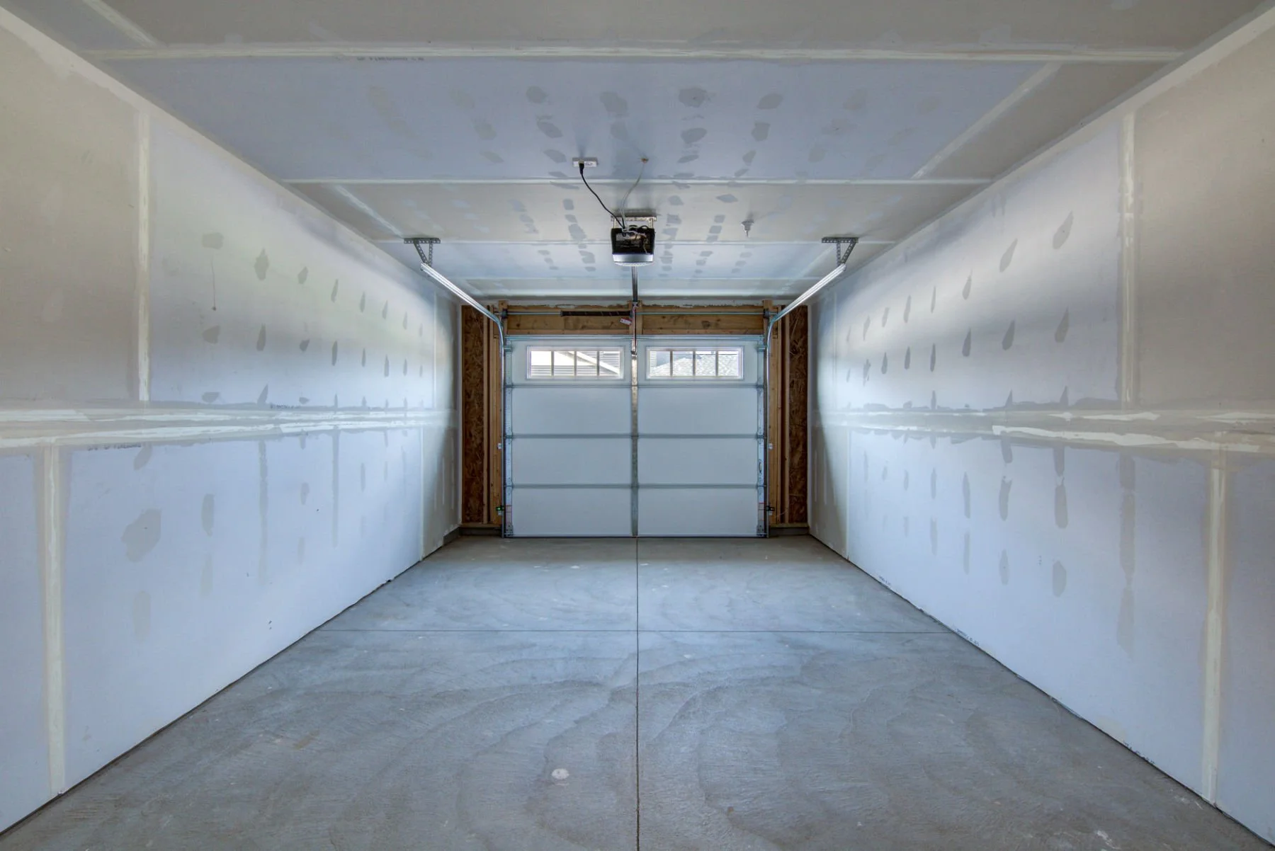Empty garage with unfinished drywall walls, concrete floor, ceiling, and garage door with windows.
