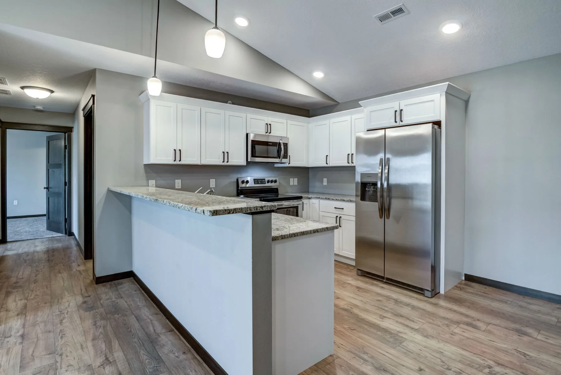 Modern kitchen with white cabinets, stainless steel refrigerator and microwave, granite countertops, and hardwood floors. Pendant lights hang above the countertop.