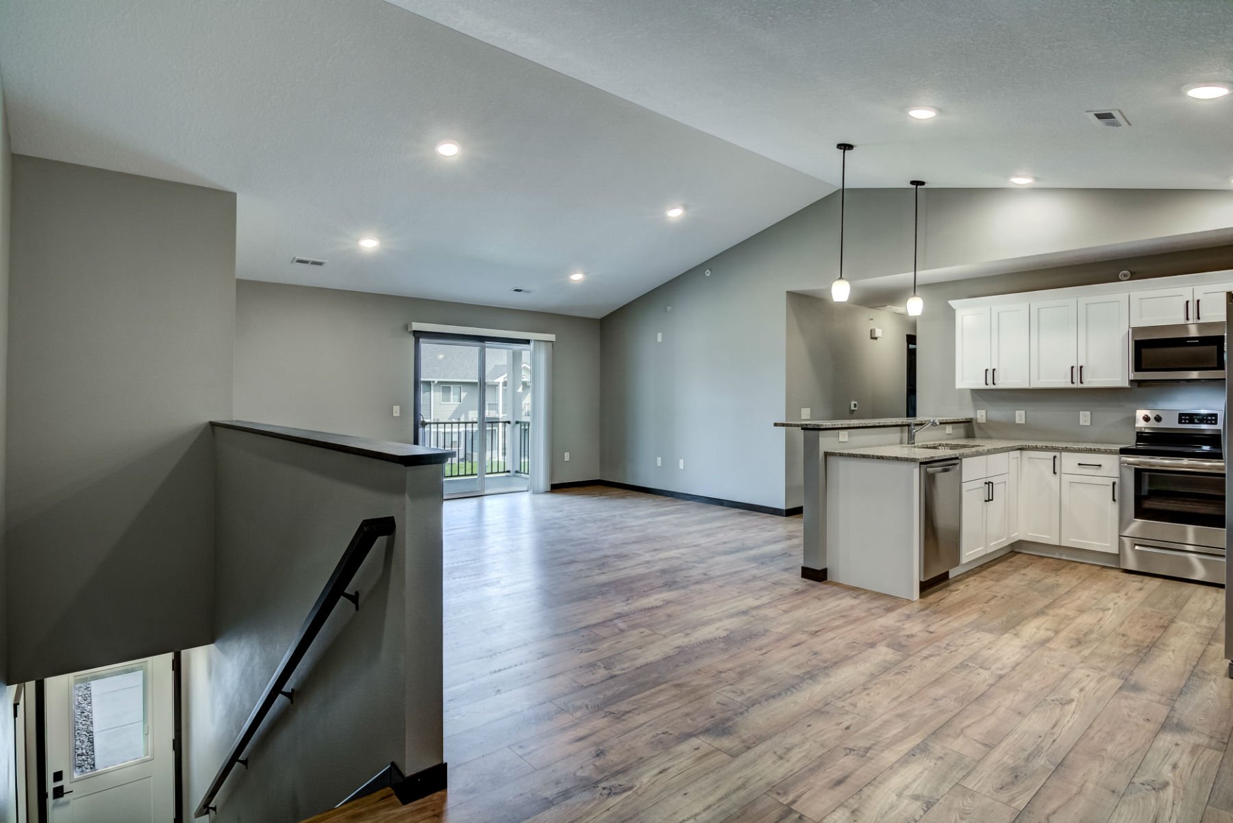 Open-concept living space with kitchen, gray walls, hardwood floors, sliding glass door leading to balcony, stainless steel appliances, white cabinets, granite countertops, and ceiling lighting fixtures.
