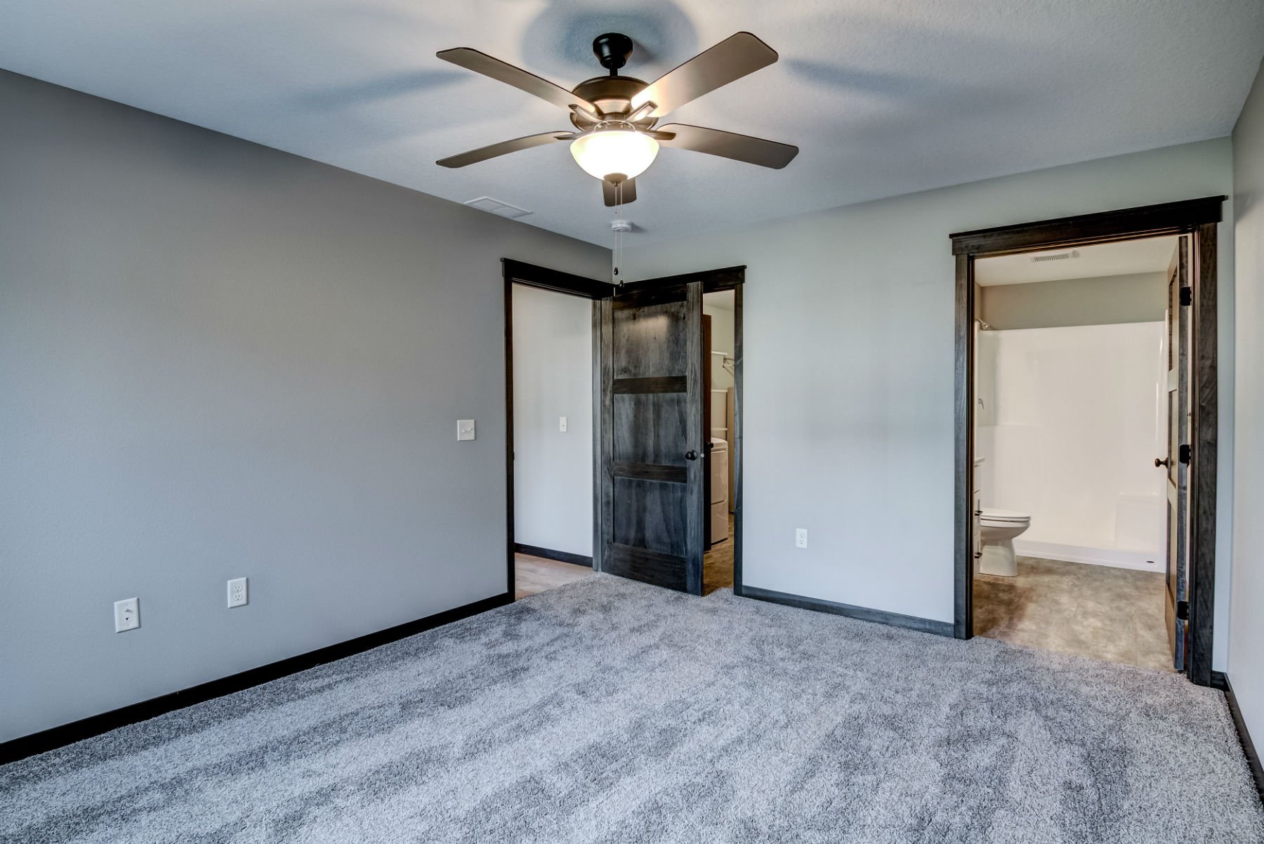 Empty bedroom with grey walls, plush grey carpet, ceiling fan with light, and two doorways leading to a bathroom and possibly a closet. The bathroom has a toilet and tiled floor.