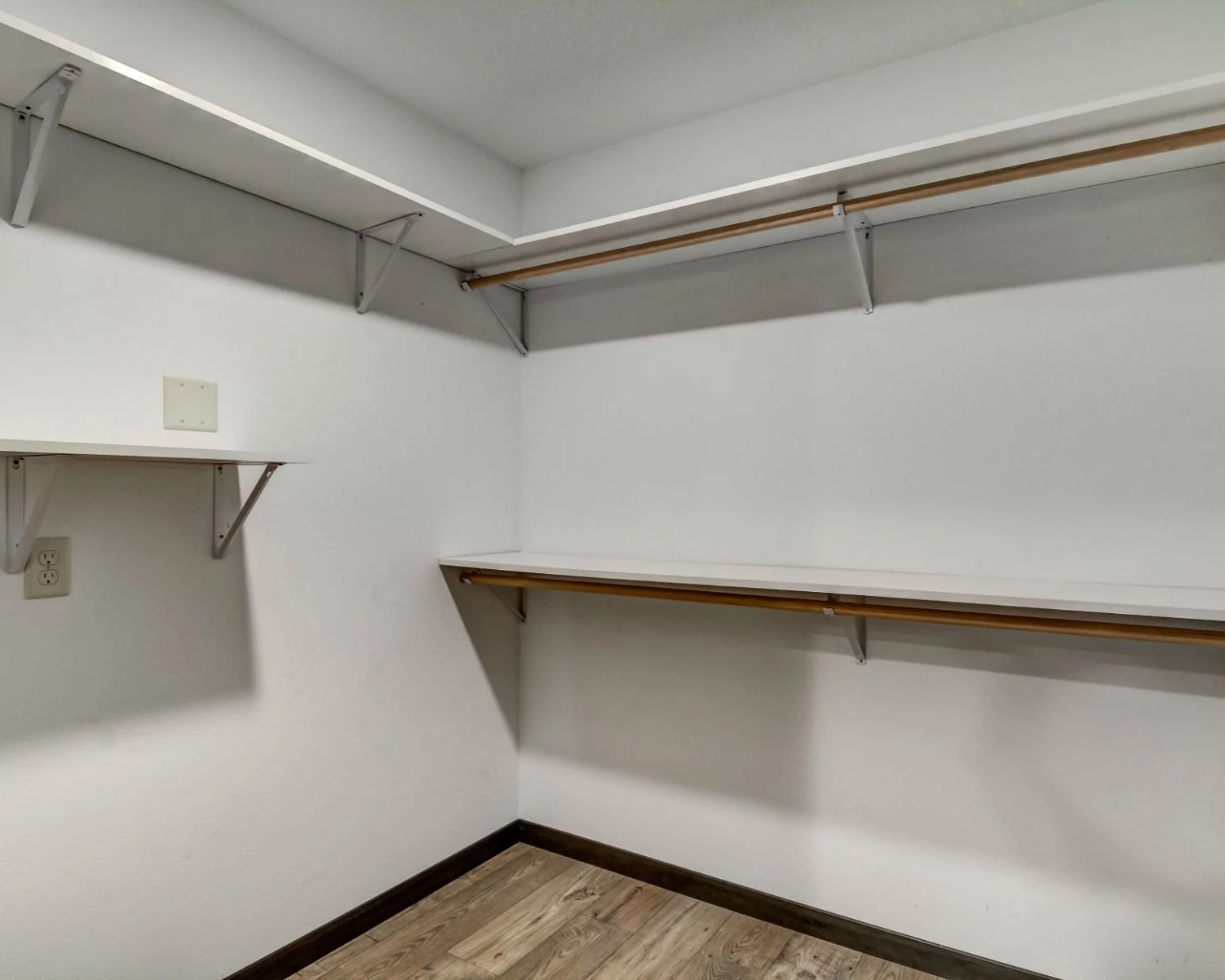 Empty walk-in closet with white shelves and wooden rods for hanging clothes, wall outlet, and wood flooring.