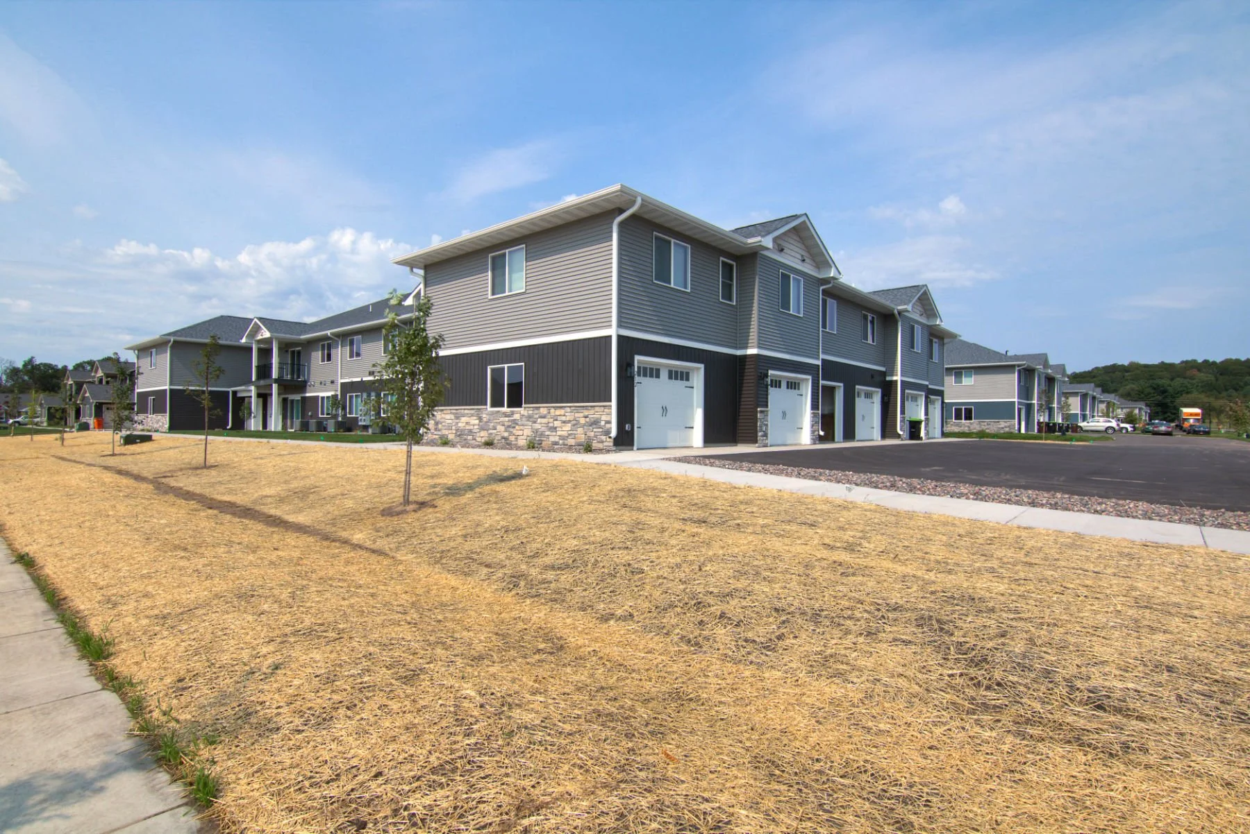 Modern apartment complex with multiple units, gray siding, stone accents, and attached garages, surrounded by a parking lot, with a grassy area and young trees in the foreground.