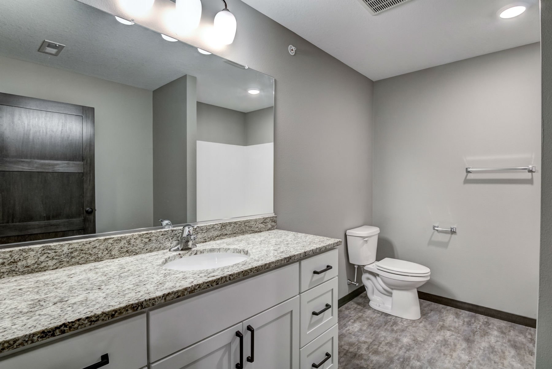 A modern bathroom with a granite countertop vanity, a large mirror, a white toilet, and gray walls with towel racks. Recessed lighting and a vent are visible on the ceiling.