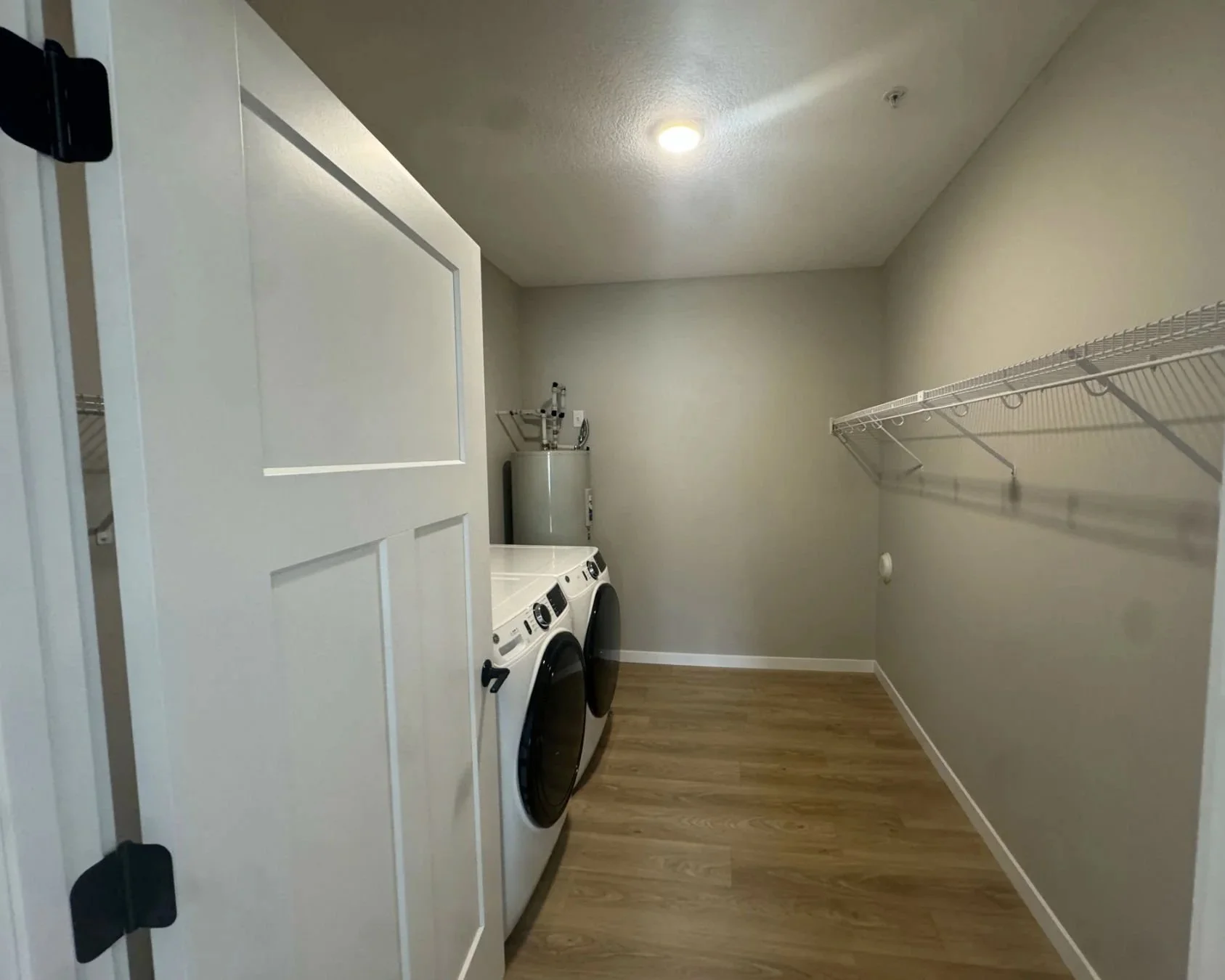 A laundry room with a washer and dryer side by side, a water heater in the corner, beige walls, a wood floor, and a wire shelf on the right wall.