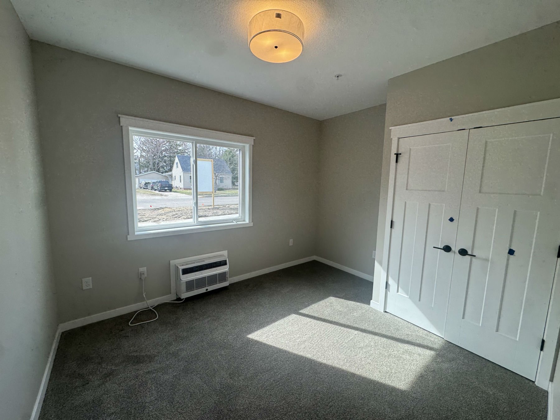 Empty bedroom with beige walls, gray carpet, a window showing a street with parked cars and houses, a white ceiling light fixture, an electric heater below the window, and a white double-door closet.
