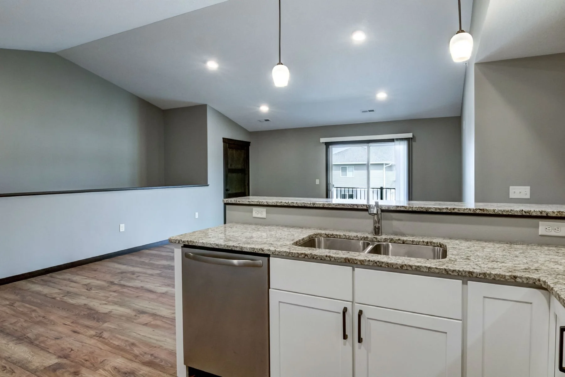 Interior view of a modern kitchen with granite countertops, white cabinets, stainless steel sink, and pendant lighting, open to a living area with wooden flooring and a sliding glass door leading to a balcony.