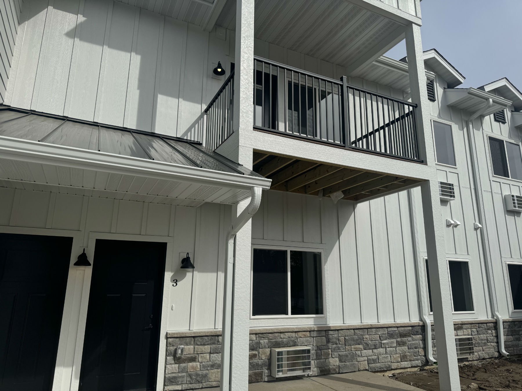Exterior view of a modern multi-story townhouse with white siding, black railings on the balcony, and a stone lower facade. The building has multiple windows, some with black shutters, and air conditioning units installed outside. There are exterior 