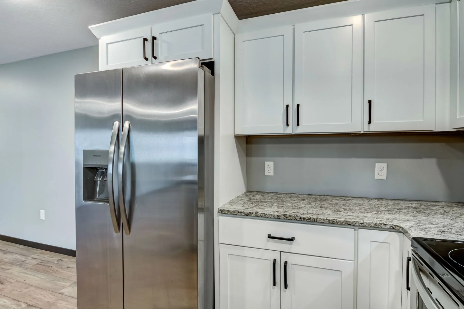 Modern kitchen with stainless steel refrigerator, white cabinets with black handles, granite countertop, and two electrical outlets on the gray wall.