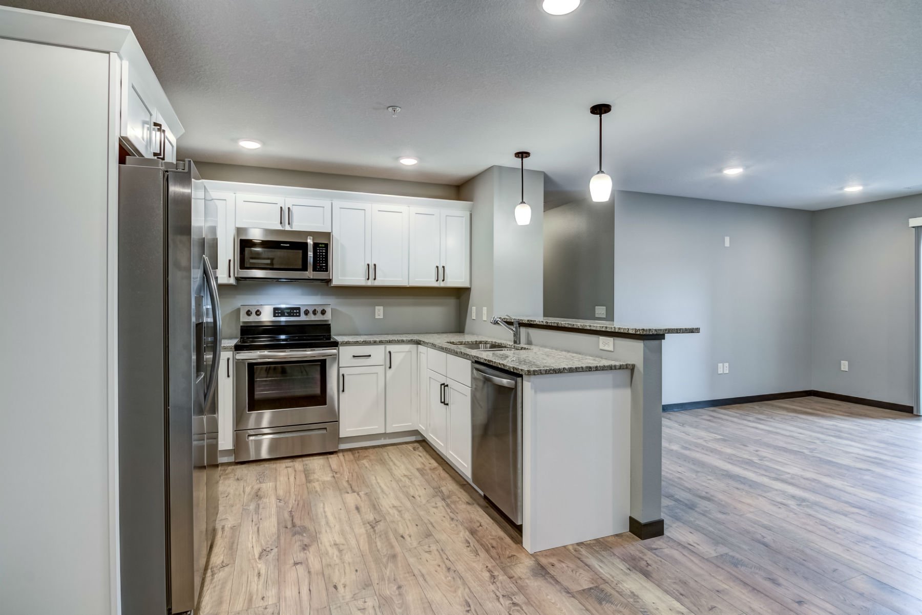 Modern kitchen with white cabinets, stainless steel appliances including a microwave, oven, refrigerator, and dishwasher. The kitchen has granite countertops, wood flooring, and is connected to an open living area with ceiling lighting and pendant li