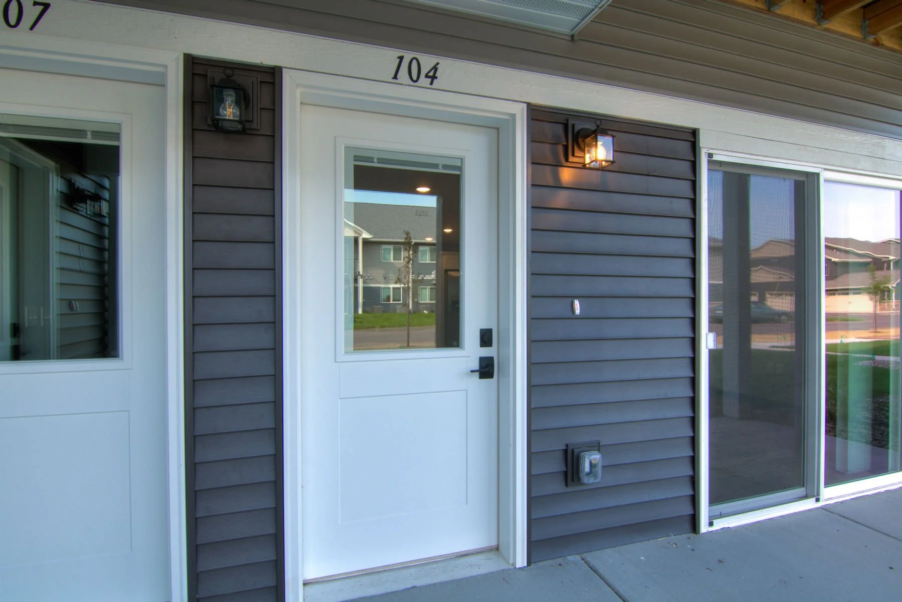 A house exterior with dark gray horizontal siding, a white front door with a large glass pane, and a sliding glass door to the right. There are outdoor wall lanterns on each side of the door. The house number 104 is above the door, and the house is r