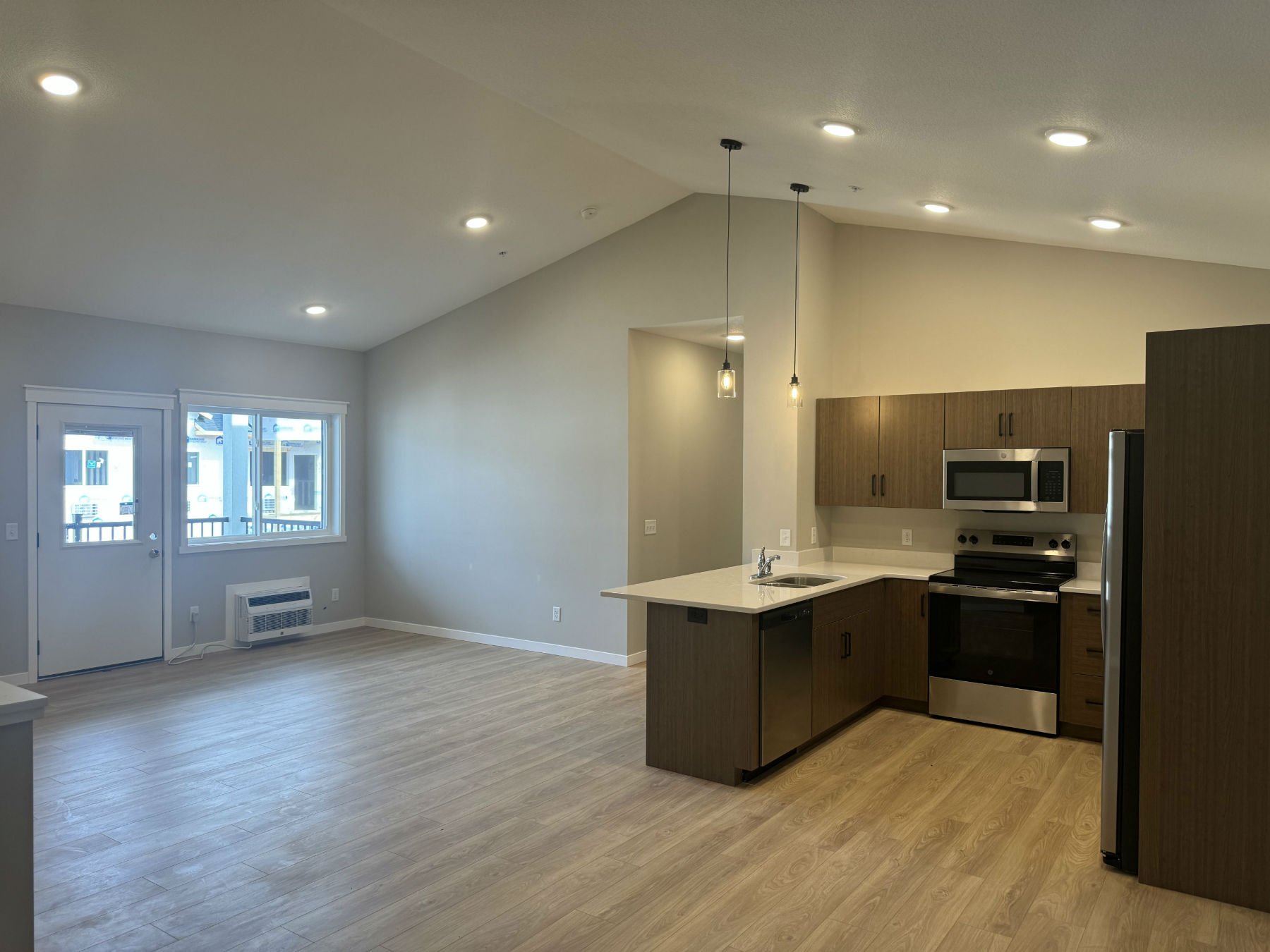 Open concept kitchen with light wood floors, modern wood cabinets, stainless steel appliances, pendant lights, white countertops, and large windows letting in natural light.