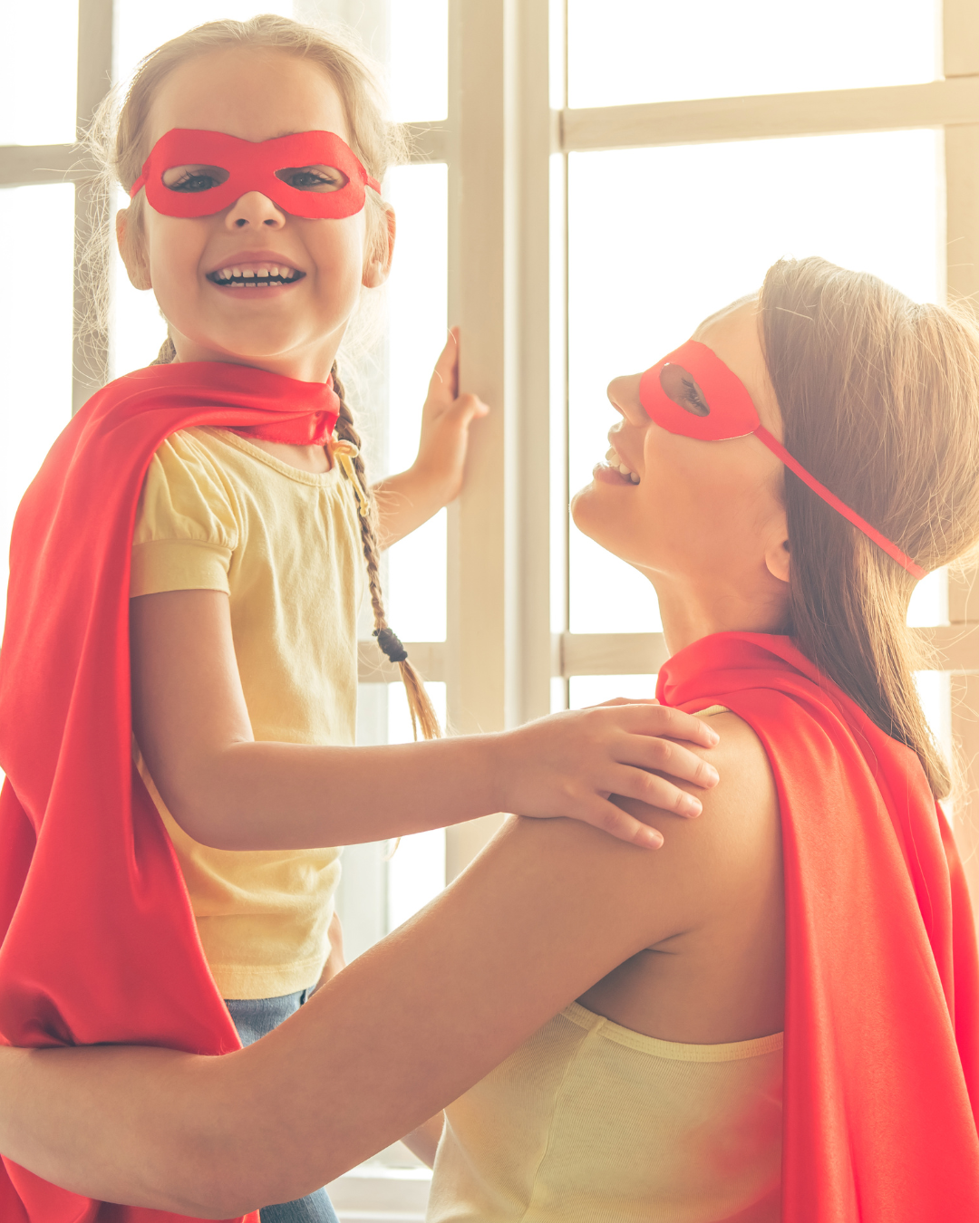 Una niña y una mujer adult mujer disfrazadas de superhéroes con máscaras y capas rojas, sonriendo y disfrutando juntas junto a una ventana brillante.
