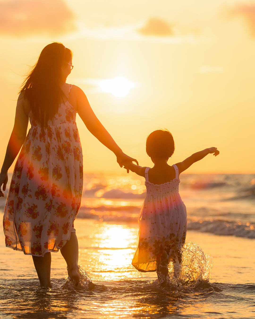 Una madre y su hija caminando por la playa durante el atardecer, sosteniéndose de las manos, con el sol brillando en el horizonte y el agua salpicando en sus pies.