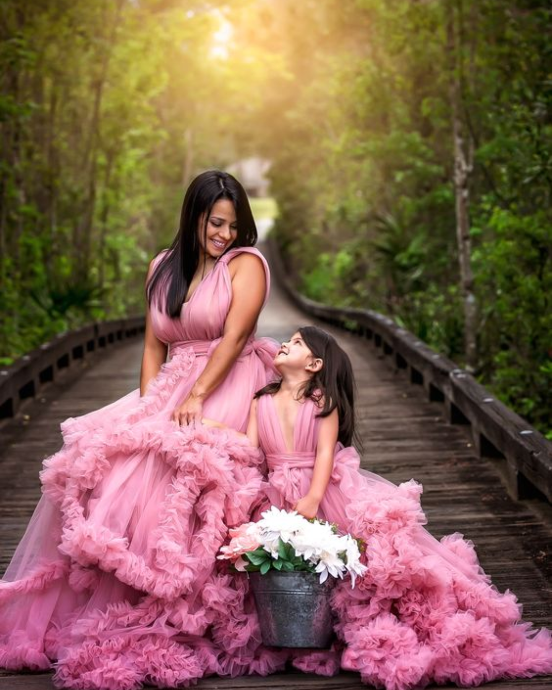 Mujer y niña con vestidos rosas en un puente de madera en un bosque, compartiendo un momento feliz con una maceta de flores blancas.