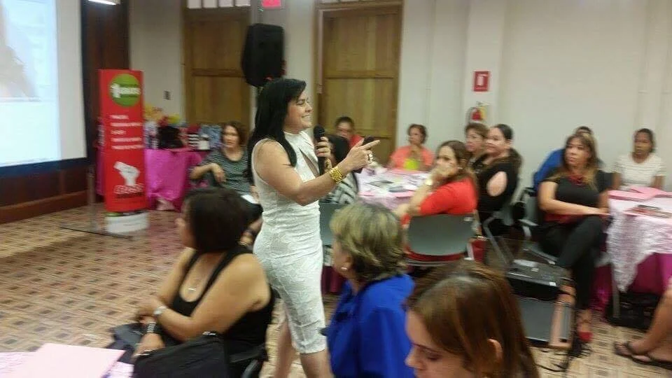 Mujer de vestido blanco hablando con micrófono frente a un grupo de mujeres en una sala de reuniones