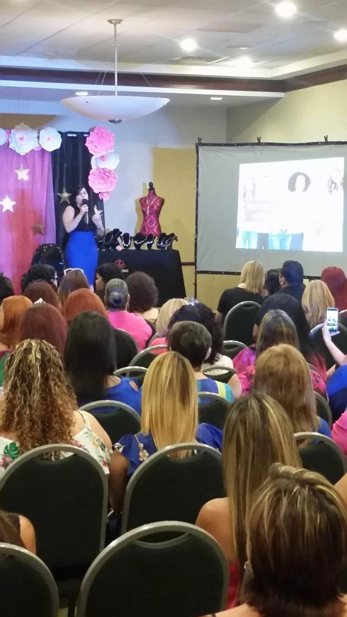 Conferencia o presentación en un salón con muchas mujeres asistiendo, decoraciones de flores y estrellas en el fondo, con una pantalla proyectando una diapositiva.
