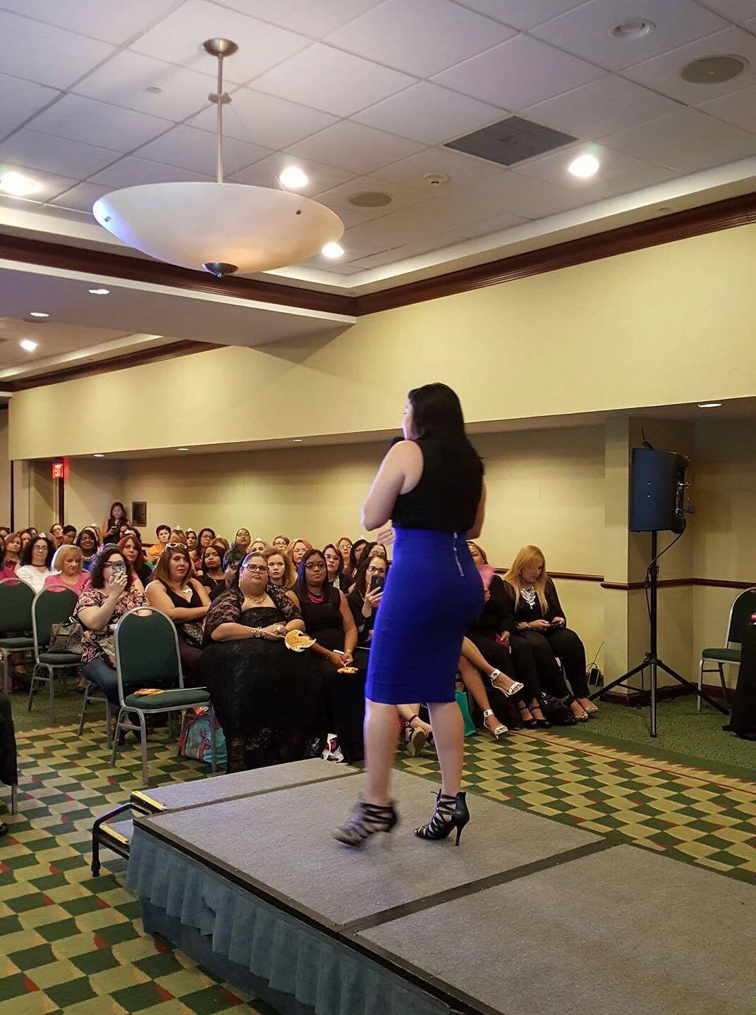 Una mujer de cabello largo de pie en un escenario, hablando frente a una audiencia de mujeres sentadas en una sala de conferencias.