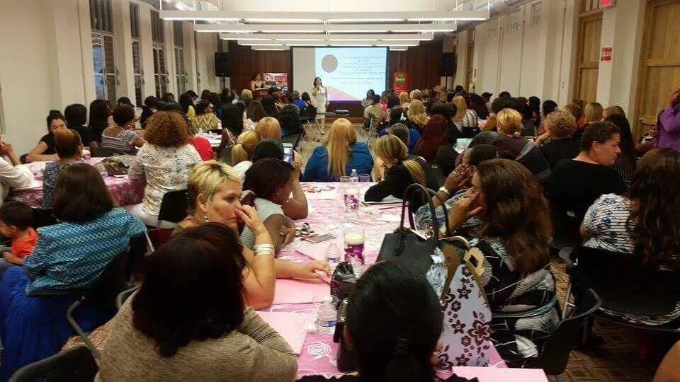 Conferencia o taller con una gran audiencia en una sala grande, asistentes sentados en mesas con manteles rosados, vista frontal del escenario y pantalla de proyector