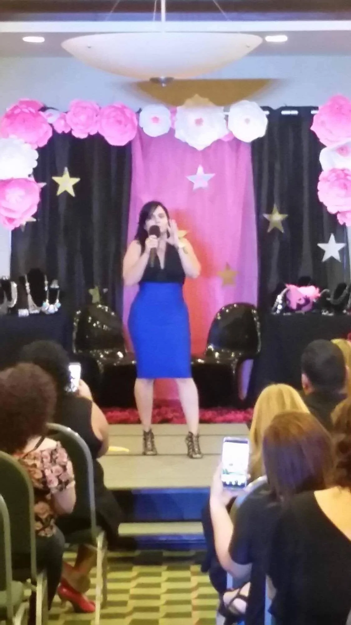 Una mujer con vestido azul y blusa negra en un escenario decorado con cortinas rosas y negras, y adornos de estrellas y flores, hablando frente a una audiencia.