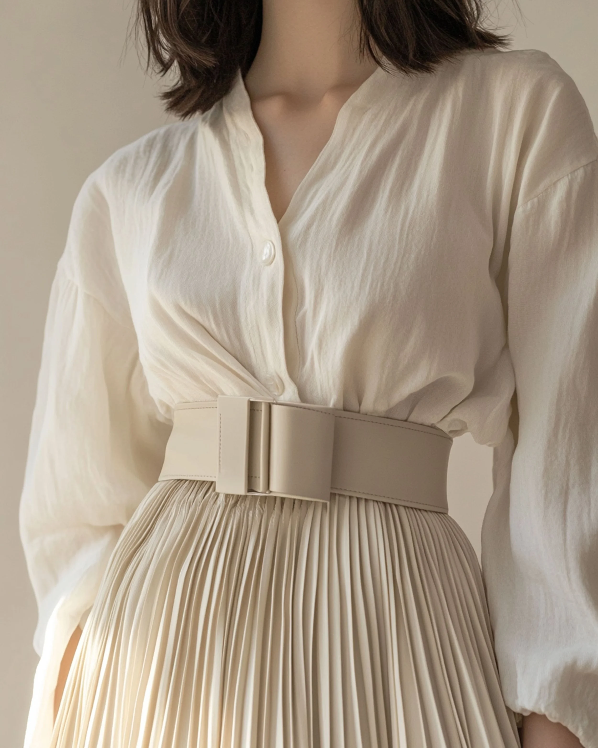 Mujer con blusa blanca de manga larga y falda plisada beige, con cinturón amplio de la misma tonalidad.
