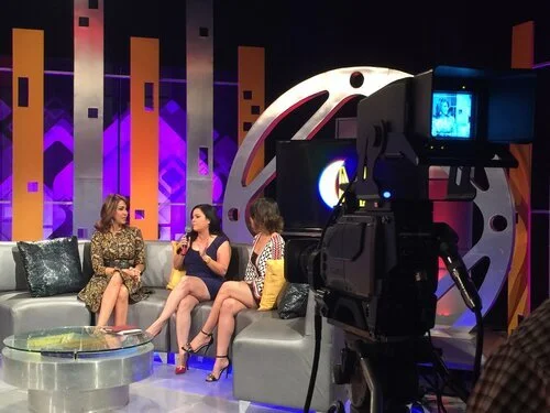 Tres mujeres sentadas en un set de televisión, con un camarógrafo grabando la entrevista, en un ambiente con decoraciones coloridas y luces moradas y naranjas.