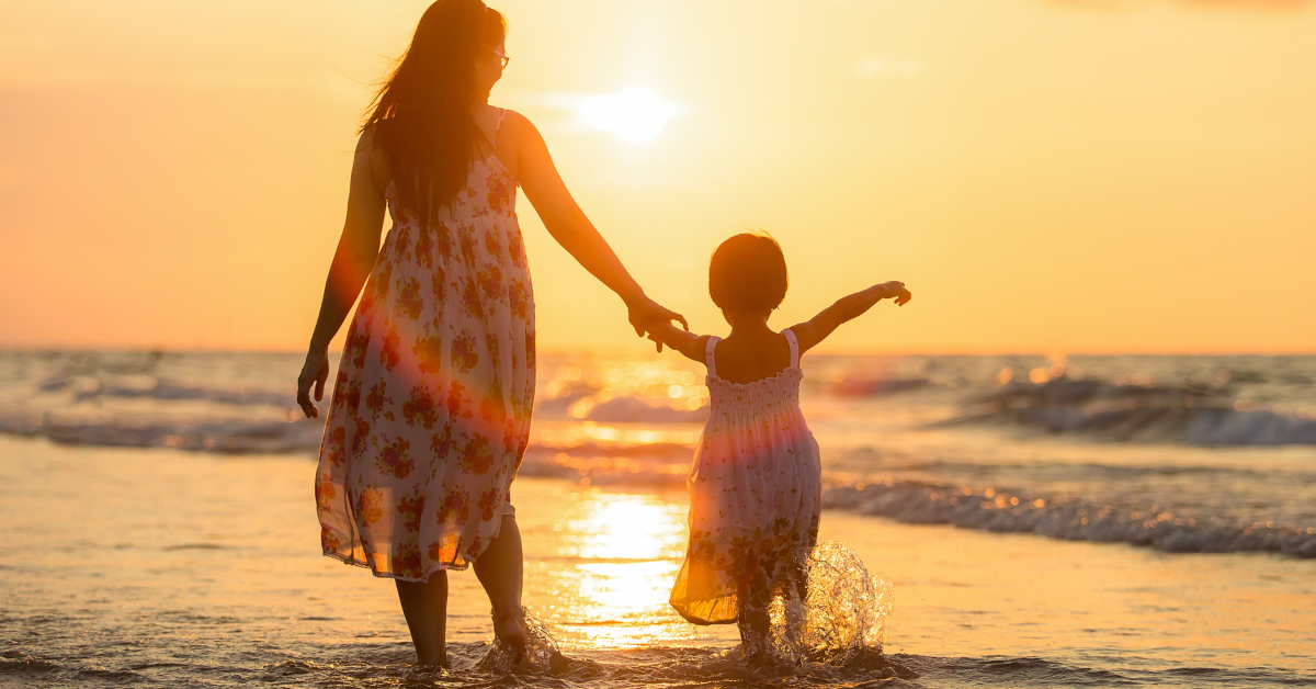 Una mujer y una niña caminando por la playa al atardecer, sosteniéndose de la mano y disfrutando del paisaje y el mar.