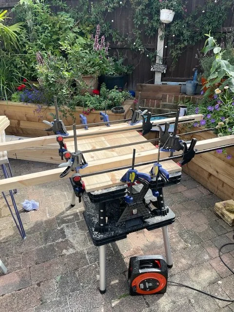 A woodworking workbench in a backyard with clamps holding a piece of wood, surrounded by plants and garden tools.