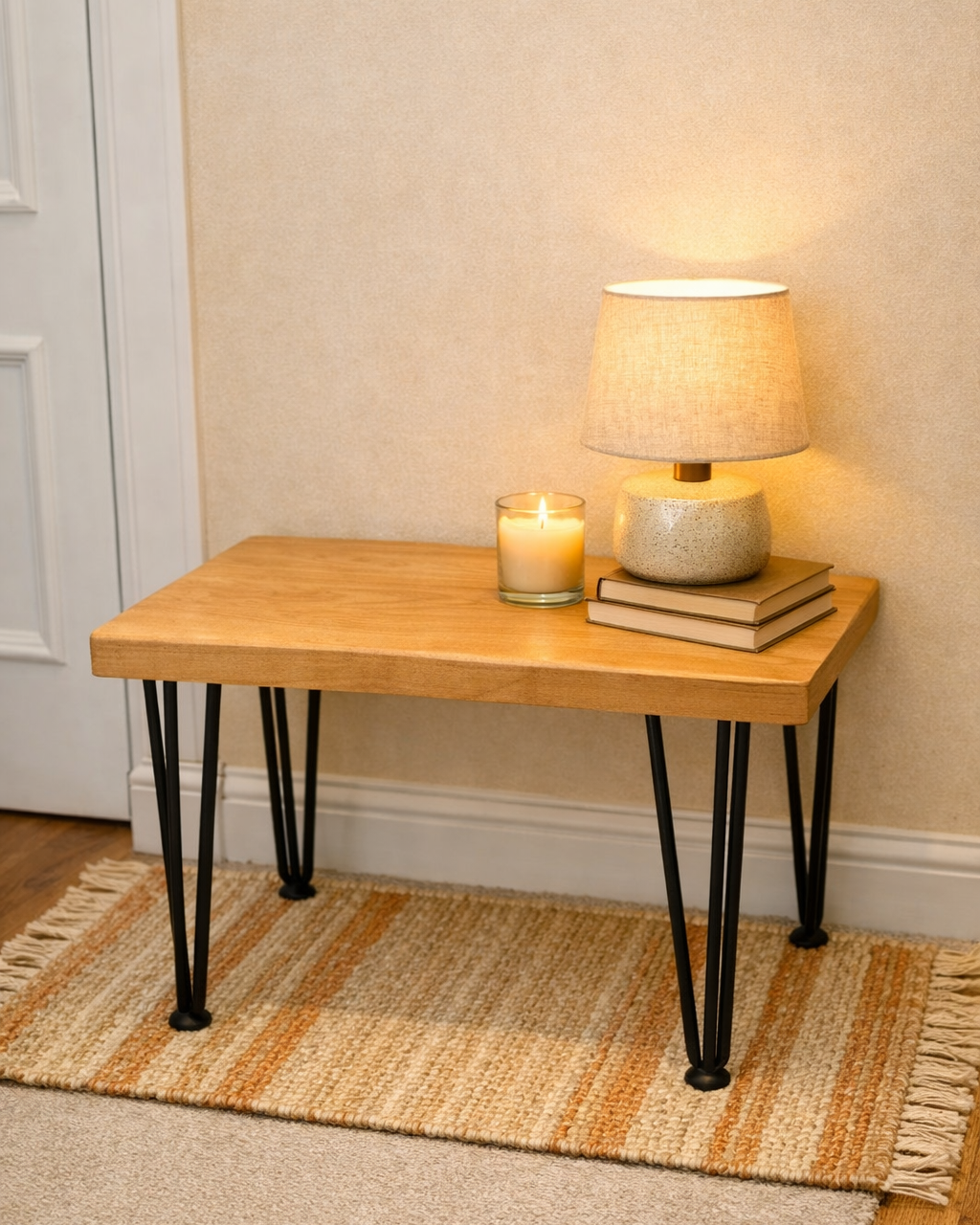 A wooden table with black hairpin legs, a lit candle in a glass holder, a table lamp with a beige shade, and three closed books on a beige and orange striped rug against a cream wall.