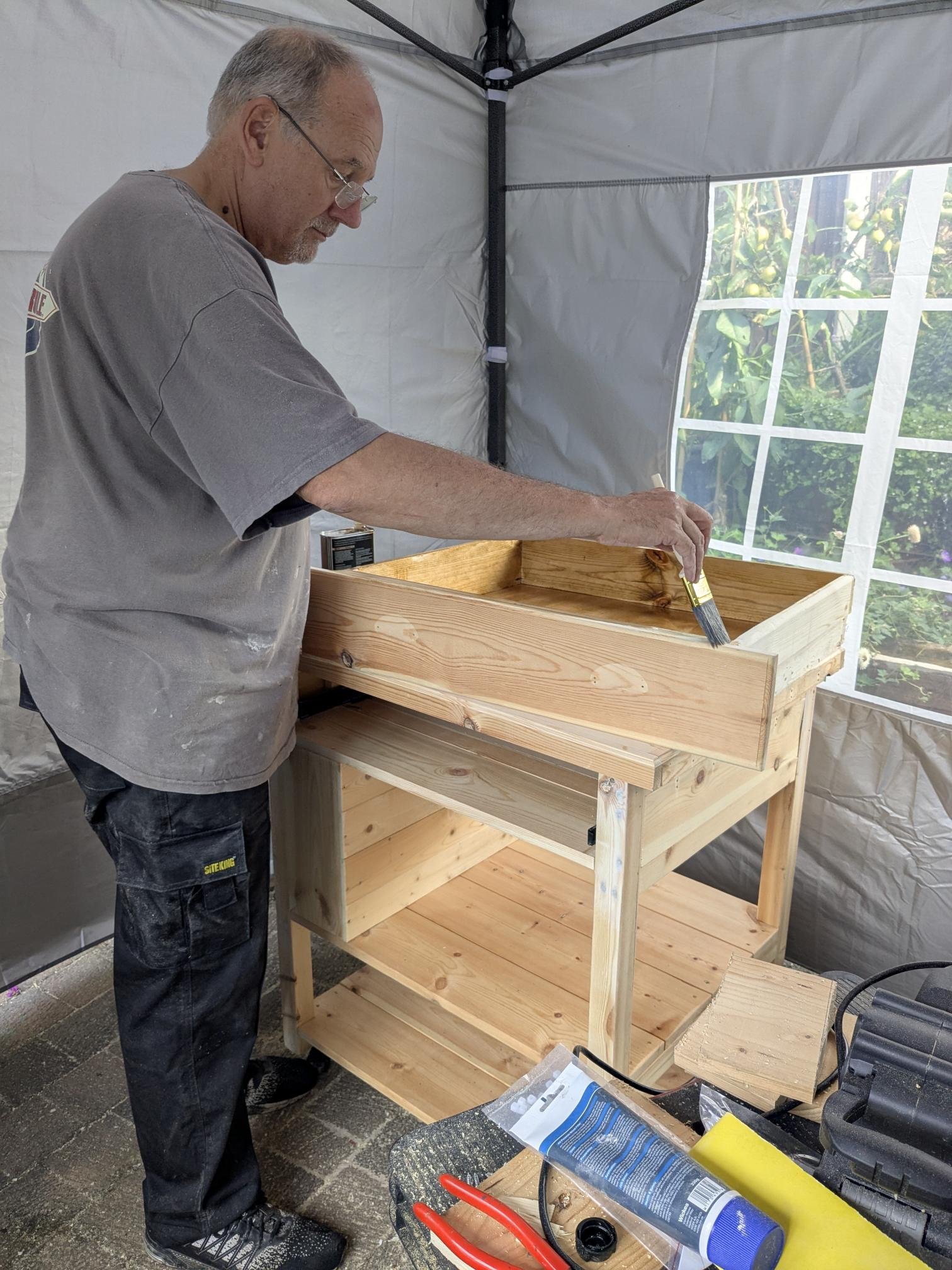 A man wearing glasses and a grey t-shirt is painting the wooden shelf of a kitchen island. The man is using a brush to apply paint or stain. There are workshop tools and supplies on the table in front of him, and a window with a view of greenery.