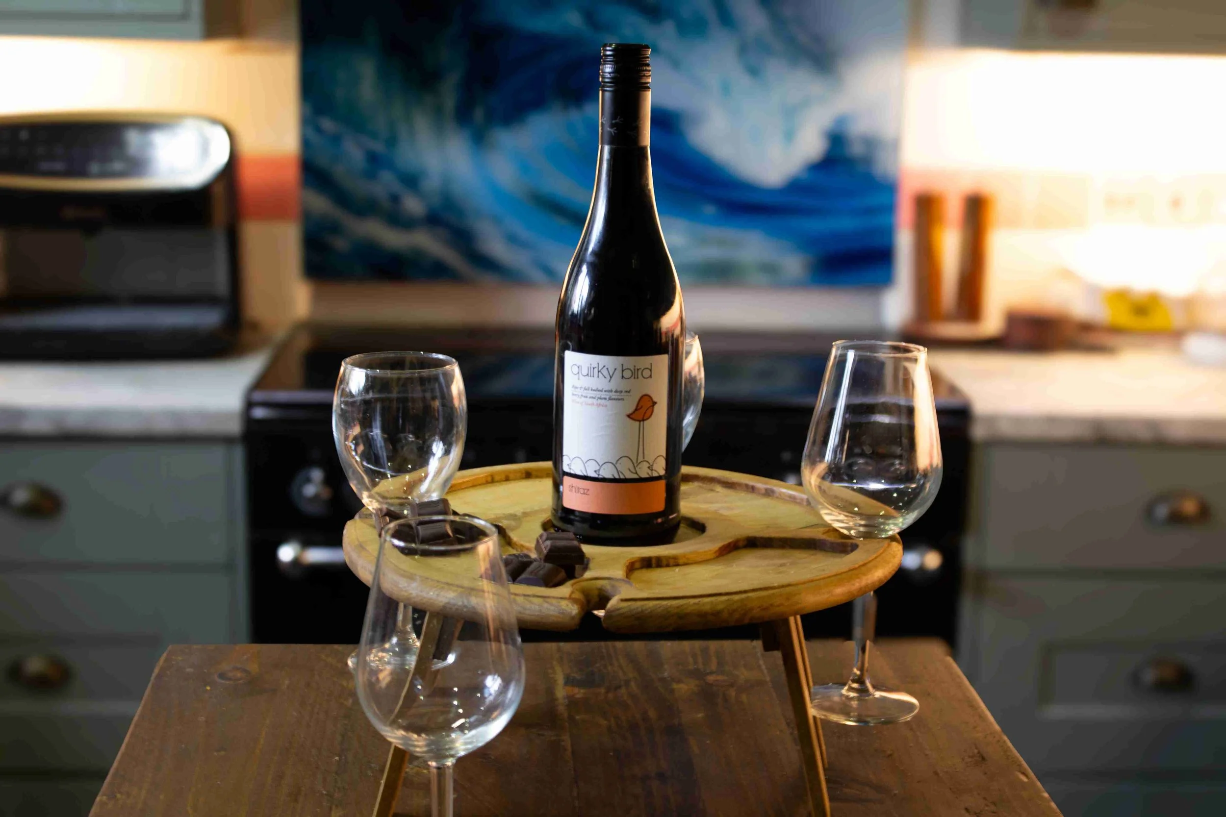 A bottle of wine labeled 'quirky bird' on a wooden table, surrounded by empty wine glasses, with a blurred kitchen background.