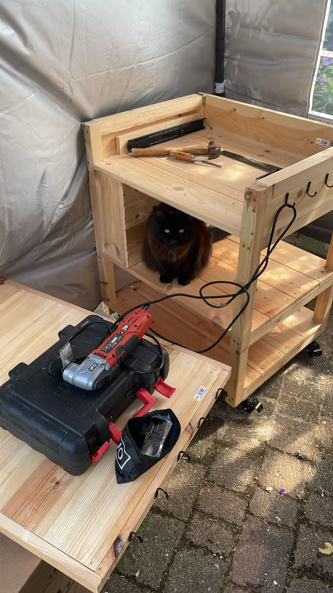 A wooden kitchen island with tools including a hammer, ruler, and power drill, and a black cat sitting underneath it. This kitchen island is being made by a carpenter and this cat is called Moose.