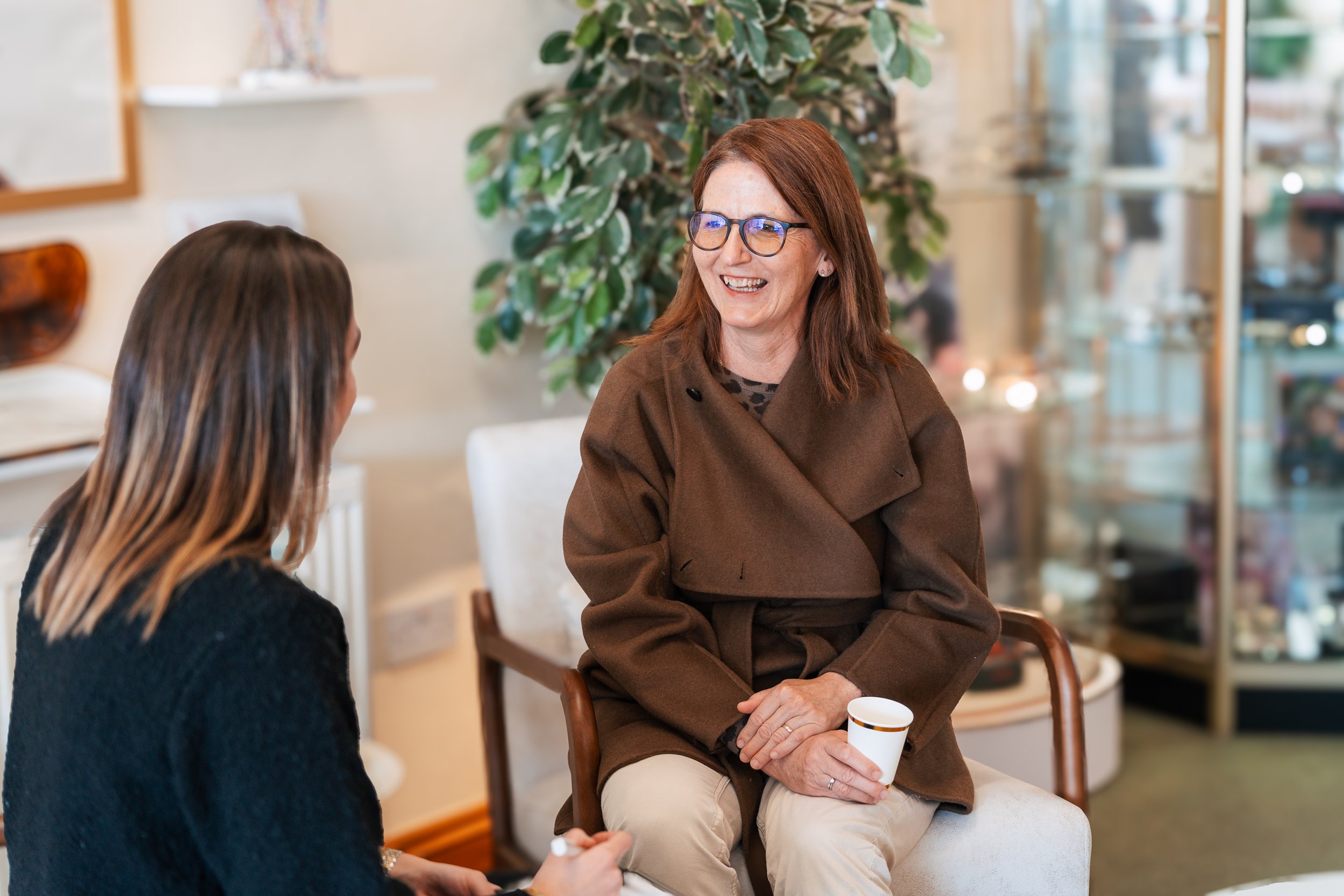 A woman with red hair and glasses is smiling while sitting and talking with a girl with long brown hair in a cozy indoor setting. The woman is wearing a brown coat, cream pants, and holding a coffee mug. There is a large green plant behind them and a glass display case in the background.